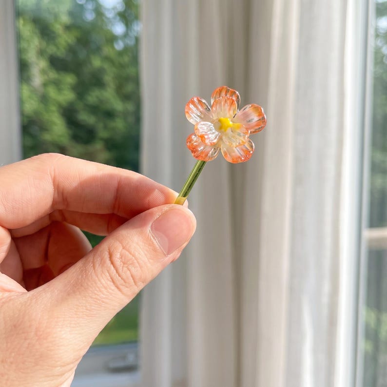 May include: A miniature, handcrafted flower with orange and white petals and a yellow centre. The flower is held by a green stem. The flower is a decorative item.