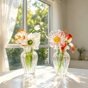 May include: Two glass vases, each containing a handcrafted flower arrangement. One vase holds orange and white flowers, the other a daisy-like flower with a yellow centre and red accents. The vases are on a white surface.