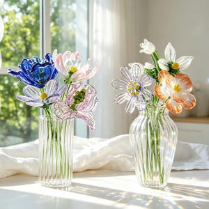 May include: Two glass vases displaying colourful glass flowers. One vase contains blue, pink, and purple blooms. The other vase holds white and orange flowers. The clear vases have vertical ridges.