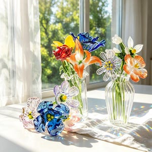 May include: A display of vibrant glass flowers in clear vases. The flowers include red roses, yellow and blue tulips, orange lilies, and white and orange daisies. The vases are arranged on a white surface near a window.