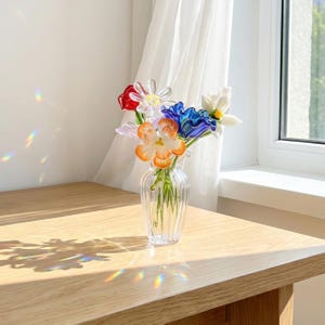 May include: A clear glass vase holding a bouquet of colourful glass flowers. The flowers are red, white, blue, and orange. The vase is on a light-coloured wooden table near a window with a sheer white curtain.