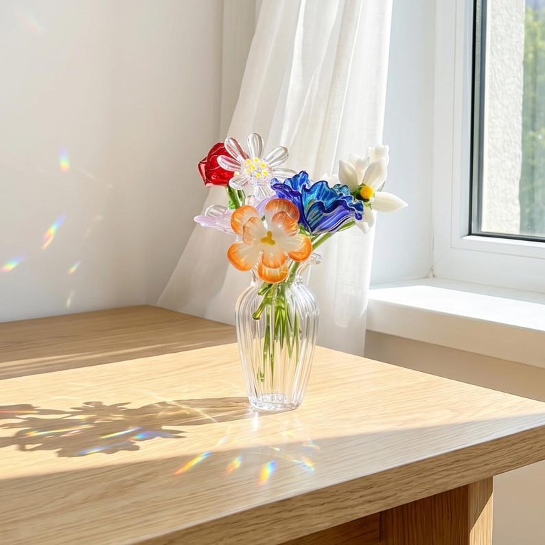 May include: A clear glass vase holding a bouquet of colourful glass flowers. The flowers are red, white, blue, and orange. The vase is on a light-coloured wooden table near a window with a sheer white curtain.