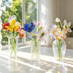 May include: Three glass vases with colourful glass flowers sit on a white table. The flowers include red, yellow, orange, blue, and purple blooms. The vases are clear and have different designs.