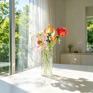 May include: A clear glass vase holding a selection of colourful glass flowers, including a red rose, orange lily, and pink and white blooms. The vase is on a white table near a window with natural light.