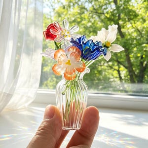 May include: A small glass vase containing a collection of colourful glass flowers. The flowers are red, white, blue, and orange. The clear, ribbed vase is held in a hand, with a blurred green backdrop visible through a window.