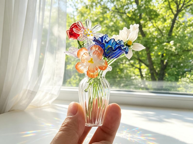 May include: A small glass vase containing a collection of colourful glass flowers. The flowers are red, white, blue, and orange. The clear, ribbed vase is held in a hand, with a blurred green backdrop visible through a window.
