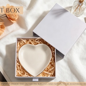 Peut inclure: Une bo&icirc;te cadeau carr&eacute;e blanche contenant un plat en c&eacute;ramique en forme de c&oelig;ur nich&eacute; dans du papier d&eacute;chiquet&eacute;. Le plat pr&eacute;sente une surface cr&egrave;me lisse avec un bord dor&eacute; fin. Le couvercle est pos&eacute; &agrave; c&ocirc;t&eacute; de la base ouverte sur un tissu blanc.