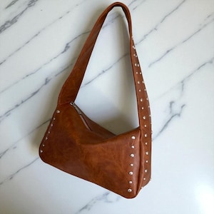 Peut inclure: Un sac à bandoulière en cuir marron avec des clous argentés le long des bords et de la bandoulière. Le sac a une fermeture éclair et une longue et fine bandoulière. Le sac est en cuir lisse et texturé.