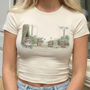 Puede incluir: Camiseta de color crema, de manga corta, con una ilustraci&oacute;n de dibujos animados de Grogu de The Mandalorian. El gr&aacute;fico muestra a Grogu en varias poses en un entorno forestal. La camiseta tiene una textura acanalada.