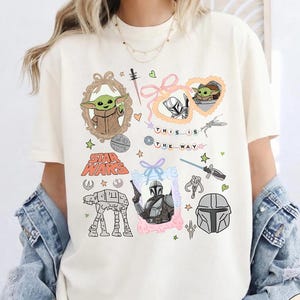 May include: Ivory-colored t-shirt featuring Star Wars-themed graphics. The design includes images of Grogu, the Mandalorian, and other iconic elements. The text "THIS IS THE WAY" is also present. The shirt is a Comfort Colors brand.
