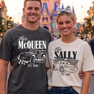 May include: Two people wearing matching t-shirts with car graphics. One shirt says "Her McQueen" and the other says "His Sally", both with "EST. 2026". The shirts are dark gray and cream-colored, respectively. The background shows a castle and a street.