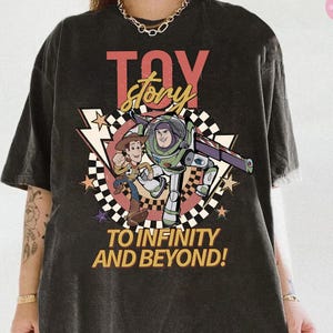 May include: Dark grey t-shirt with a Toy Story graphic. The design features the words "Toy Story" in red and yellow, and the phrase "To Infinity and Beyond!" The shirt shows Woody and Buzz Lightyear.