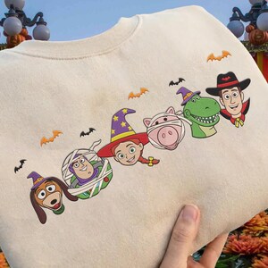May include: Cream-colored sweatshirt featuring embroidered Halloween-themed Toy Story characters. The design includes Slinky Dog, Buzz Lightyear, Jessie, a pig, Rex, and Woody, each in a Halloween costume, with orange bat accents.