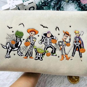 May include: A cream-colored sweatshirt featuring an embroidered graphic of five cartoon characters dressed in skeleton costumes for Halloween. Each character holds a small orange pumpkin, surrounded by black bats, stars, and spider webs.