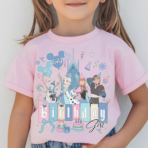 May include: A light pink t-shirt featuring a colorful graphic of characters from a popular animated movie, including a castle, reindeer, and a snowman. The shirt has the text "Birthday Girl" and a name on a balloon.