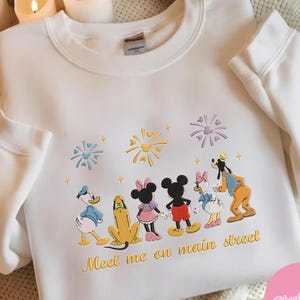 May include: White sweatshirt featuring a colorful embroidered design of Disney characters, including Mickey and Minnie Mouse, Donald Duck, and Goofy. The phrase "Meet me on main street" is embroidered in gold. The sweatshirt has long sleeves and a crew neck.