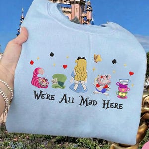 Puede incluir: Sudadera azul claro con un dise&ntilde;o bordado de personajes de Alicia en el pa&iacute;s de las maravillas. El dise&ntilde;o incluye el Gato de Cheshire, el sombrero del Sombrerero Loco, Alicia, el Sombrerero Loco y tazas de t&eacute;. El texto "We're All Mad Here" est&aacute; bordado.
