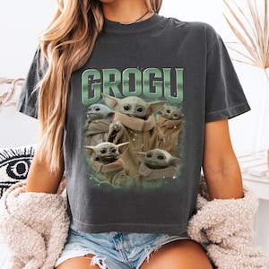 May include: Dark gray t-shirt featuring the word "GROGU" in green block letters above a graphic of Grogu from The Mandalorian. The graphic shows Grogu in various poses. The shirt is a crew neck.