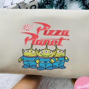 May include: A beige clutch bag with a red embroidered 'Pizza Planet' logo and a planet graphic. Below the logo are three green aliens in blue jumpsuits. The bag is rectangular in shape.