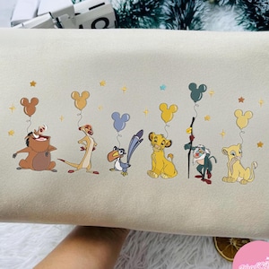 K&ouml;nnte beinhalten: Cremefarbener Kissenbezug mit gestickten Charakteren aus Der K&ouml;nig der L&ouml;wen, darunter Timon, Pumbaa, Simba und Zazu. Jeder Charakter h&auml;lt einen Mickey Mouse-f&ouml;rmigen Ballon. Goldene Sterne und Mickey-Luftballons sind &uuml;ber das Design verstreut.