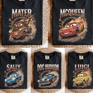 May include: Black t-shirts featuring characters from the movie "Cars". Each shirt displays a different character's name: Mater, McQueen, Sally, Doc Hudson, and Luigi, along with an illustration of the character. The designs have a vintage, distressed look.