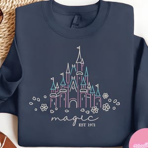 May include: Navy blue sweatshirt featuring a line drawing of a castle in pink and blue, with the word "magic" and "EST 1971" in white script. The design includes floral accents.