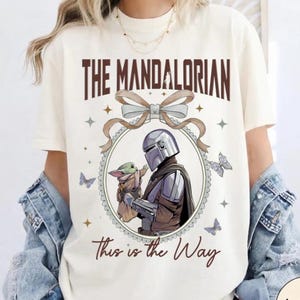 May include: Ivory t-shirt with a graphic of The Mandalorian and Grogu. The text "The Mandalorian" is above the image, and "This is the Way" is below. The design includes a bow, stars, and butterflies. The t-shirt is ivory in colour.