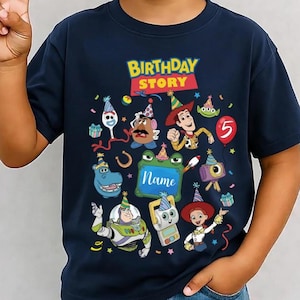 May include: A navy blue short-sleeve t-shirt featuring a colourful graphic with the text "BIRTHDAY STORY" and "Name" inside a green frame. The design includes various cartoon characters wearing party hats, a red balloon with the number 5, and confetti.