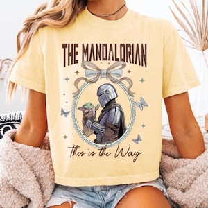 May include: Yellow t-shirt featuring a graphic from The Mandalorian. The design includes the text "THE MANDALORIAN" above an illustration of the Mandalorian and Grogu, with the phrase "This is the Way" below. Butterflies and stars surround the central image.