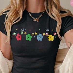 Puede incluir: Top corto negro con un dise&ntilde;o bordado de cuatro personajes de dibujos animados en rojo, verde, azul y amarillo. Cada personaje sostiene una seta y est&aacute; conectado por estrellas. La camiseta tiene mangas cortas y cuello redondo.