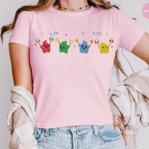 Puede incluir: Camiseta rosa claro con un dise&ntilde;o de dibujos animados. El dise&ntilde;o incluye cuatro personajes en forma de estrella en rojo, verde, azul y amarillo, cada uno sosteniendo una estrella y una seta. La camiseta es de material suave.