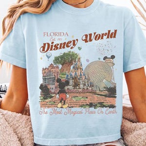 May include: Pale blue t-shirt featuring a retro Disney World design. The graphic includes the text "Florida Disney World" and "The Most Magical Place On Earth" with illustrations of Mickey Mouse and park attractions.