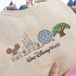Peut inclure: Sweat-shirt beige avec broderie Walt Disney World. Il pr&eacute;sente une t&ecirc;te de Mickey Mouse marron, un ch&acirc;teau rose et bleu, une sph&egrave;re bleue et blanche et un arbre vert. Les mots "Walt Disney World" sont brod&eacute;s en noir.