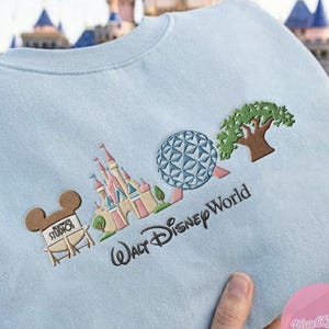 Peut inclure: Sweat-shirt bleu clair avec broderie d'ic&ocirc;nes de Disney World : ch&acirc;teau, Spaceship Earth et arbre. Le texte "Walt Disney World" est brod&eacute; en dessous. Comprend &eacute;galement une t&ecirc;te de Mickey Mouse avec le texte "Hollywood Studios".
