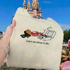 May include: Cream-colored sweatshirt featuring a cartoon character lying next to a record player. The character is wearing a red and white outfit. The text "Leave me alone to die" is printed below the graphic.