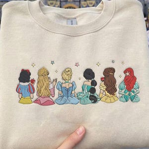 May include: Beige sweatshirt featuring an embroidered design of six cartoon princesses. The princesses are depicted from the back, each with unique hairstyles and dresses in various colors. The sweatshirt is held up in front of a castle.