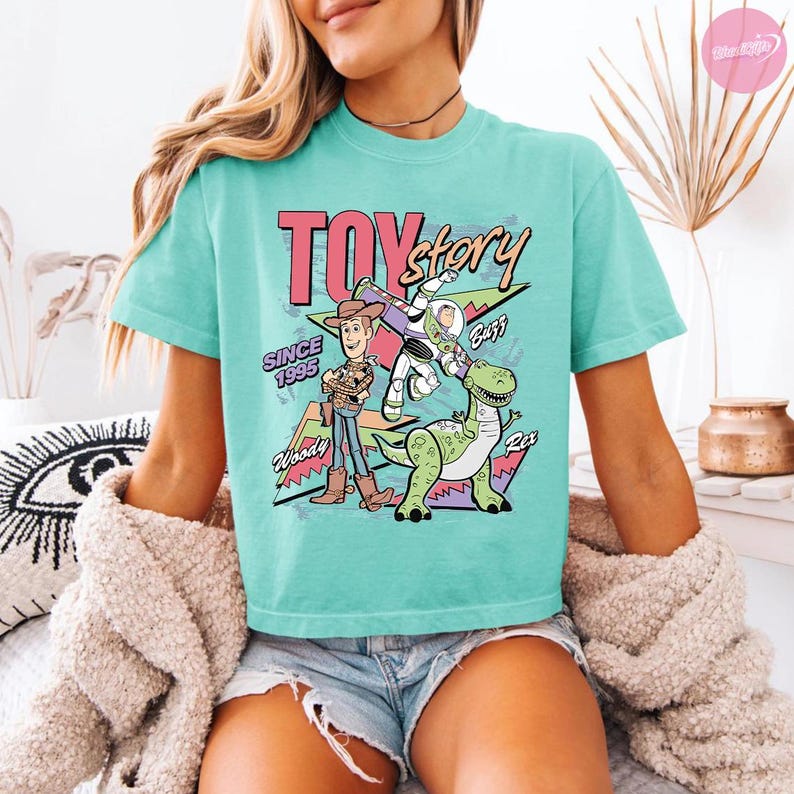 Op de afbeelding: Een turquoise t-shirt met de tekst "TOY STORY" in roze en zwart, en illustraties van Woody, Buzz Lightyear en Rex. Het shirt heeft ook de tekst "SINCE 1995".