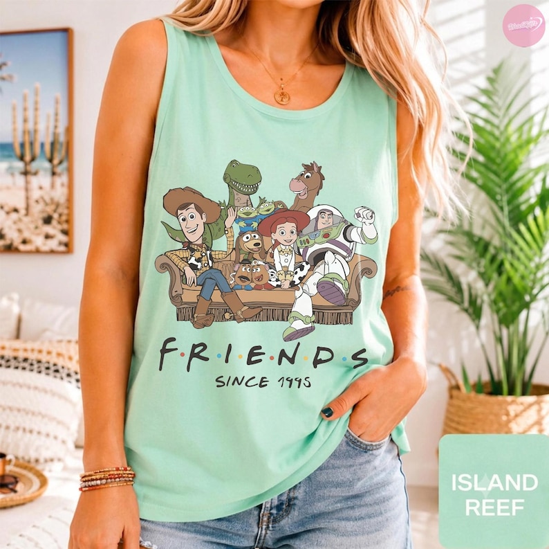 May include: A light green sleeveless tank top featuring a graphic print of various animated characters sitting on a couch. Below the characters, the text reads "FRIENDS" with colored dots between the letters and "SINCE 1995" printed underneath.