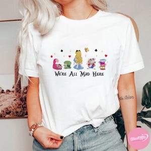 Puede incluir: Camiseta blanca con una colorida ilustraci&oacute;n de personajes de Alicia en el pa&iacute;s de las maravillas, incluyendo el Gato de Cheshire, el Sombrerero Loco, Alicia y la Liebre de Marzo. El texto "We're All Mad Here" est&aacute; impreso debajo.