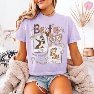 Puede incluir: Una camiseta de color lila claro con la palabra "Besties" en escritura marr&oacute;n. La camiseta presenta im&aacute;genes de dibujos animados de Chip y Chop, con los nombres "chip" y "dale" debajo. Elementos decorativos incluyen corazones y estrellas.