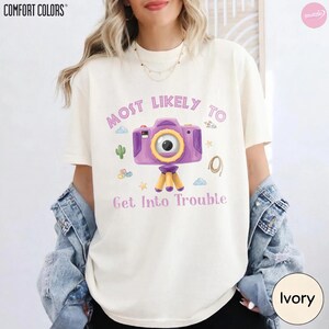 Op de afbeelding: Een ivoorkleurig T-shirt met korte mouwen en een afbeelding van een paarse camera op een statief. De tekst "MOST LIKELY TO" staat boven de camera en "Get Into Trouble" eronder, omringd door kleine illustraties van een cactus, wolken en sterren.