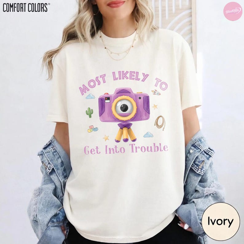 Op de afbeelding: Een ivoorkleurig T-shirt met korte mouwen en een afbeelding van een paarse camera op een statief. De tekst "MOST LIKELY TO" staat boven de camera en "Get Into Trouble" eronder, omringd door kleine illustraties van een cactus, wolken en sterren.