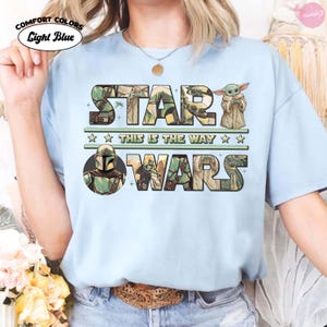 Puede incluir: Camiseta azul claro con las palabras "STAR WARS" en un dise&ntilde;o verde y marr&oacute;n, con personajes e im&aacute;genes de la franquicia Star Wars. Tambi&eacute;n se incluye el texto "THIS IS THE WAY".
