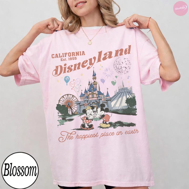 May include: A pale pink t-shirt with a Disneyland graphic. The design includes a castle, Ferris wheel, and Mickey and Minnie Mouse. Text on the shirt reads "CALIFORNIA Est. 1955 Disneyland" and "The happiest place on earth."