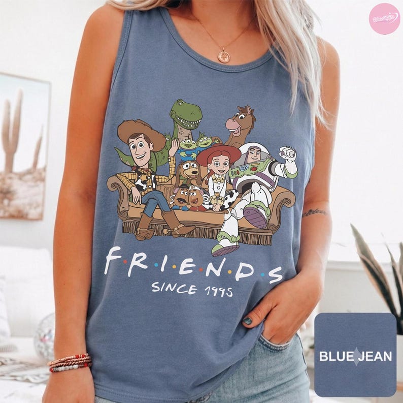 May include: A blue denim-colored tank top featuring a graphic print of Toy Story characters sitting on a couch. The text below the characters reads "FRIENDS" with colored dots between the letters and "SINCE 1995" printed underneath in white.