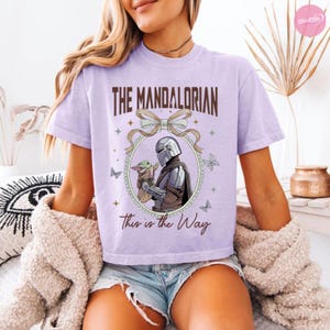 May include: A light purple t-shirt featuring the text "The Mandalorian" in brown lettering above an oval illustration of the Mandalorian and Grogu. The phrase "This is the Way" is printed below the illustration. Butterflies and stars decorate the design.