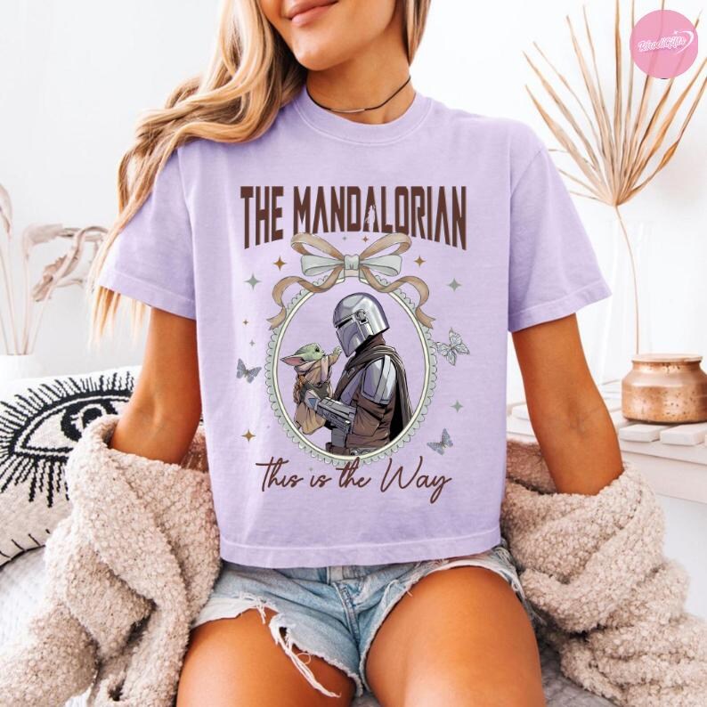 May include: A light purple t-shirt featuring the text "The Mandalorian" in brown lettering above an oval illustration of the Mandalorian and Grogu. The phrase "This is the Way" is printed below the illustration. Butterflies and stars decorate the design.