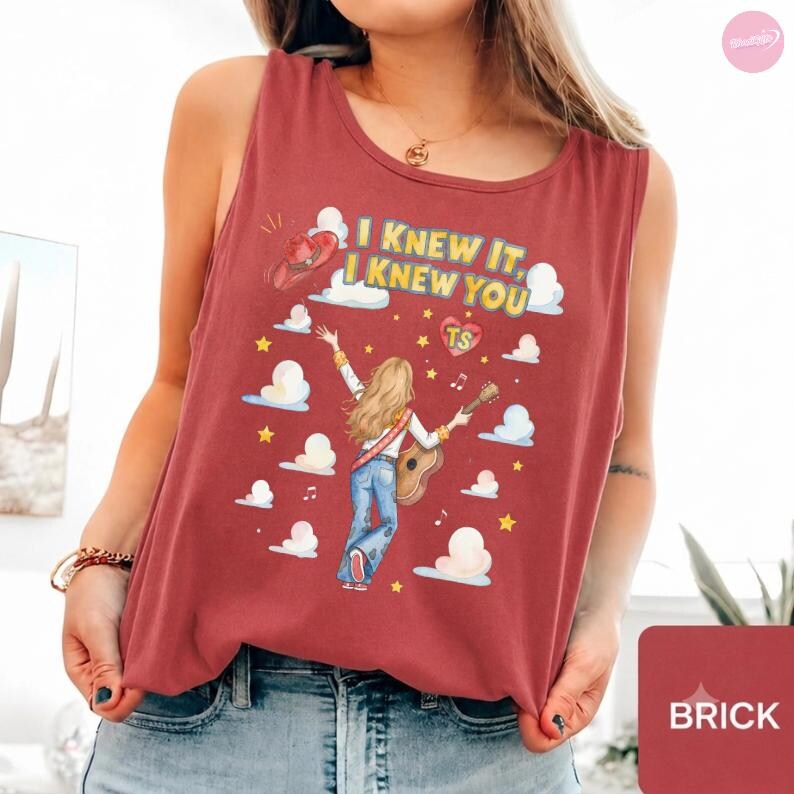 May include: A brick red sleeveless tank top featuring a graphic print of a person with long hair playing an acoustic guitar. The text reads "I KNEW IT, I KNEW YOU" with a heart containing "TS", surrounded by clouds, stars, and a cowboy hat.