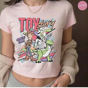 Op de afbeelding: Lichtroze T-shirt met de tekst "Toy Story" en de figuren Woody, Buzz Lightyear en Rex de dinosaurus. Het shirt bevat ook de tekst "Since 1995" in een retrostijl.