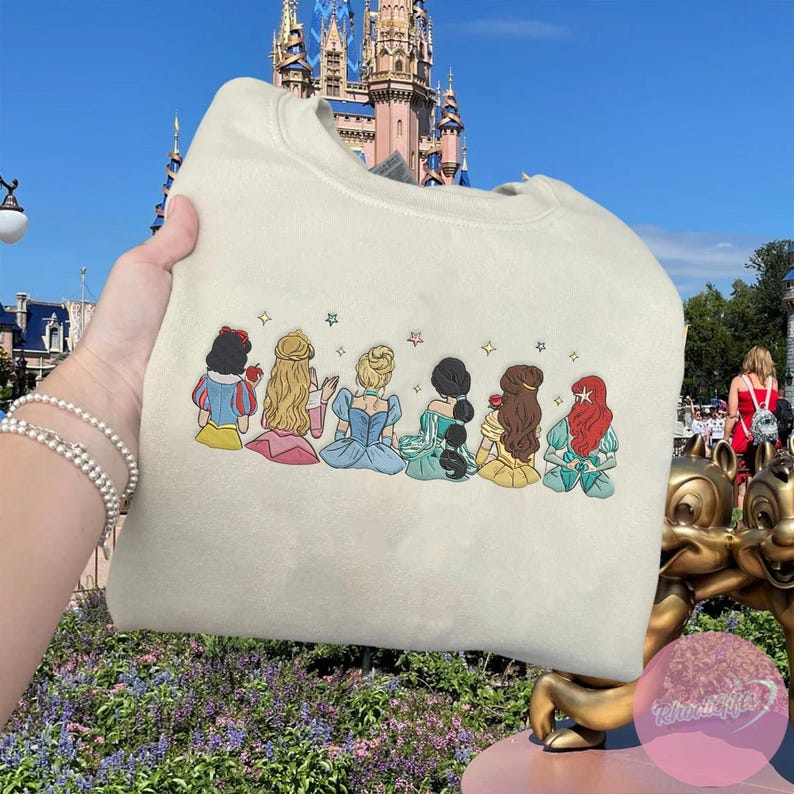 May include: Cream-colored sweatshirt featuring an embroidered design of seven cartoon princesses. Each princess is depicted from the back, with details like hair color and dress style. The design includes stars above each princess.